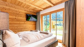 Quarto com painéis de madeira, uma cama, televisão na parede e uma janela grande com cortinas.