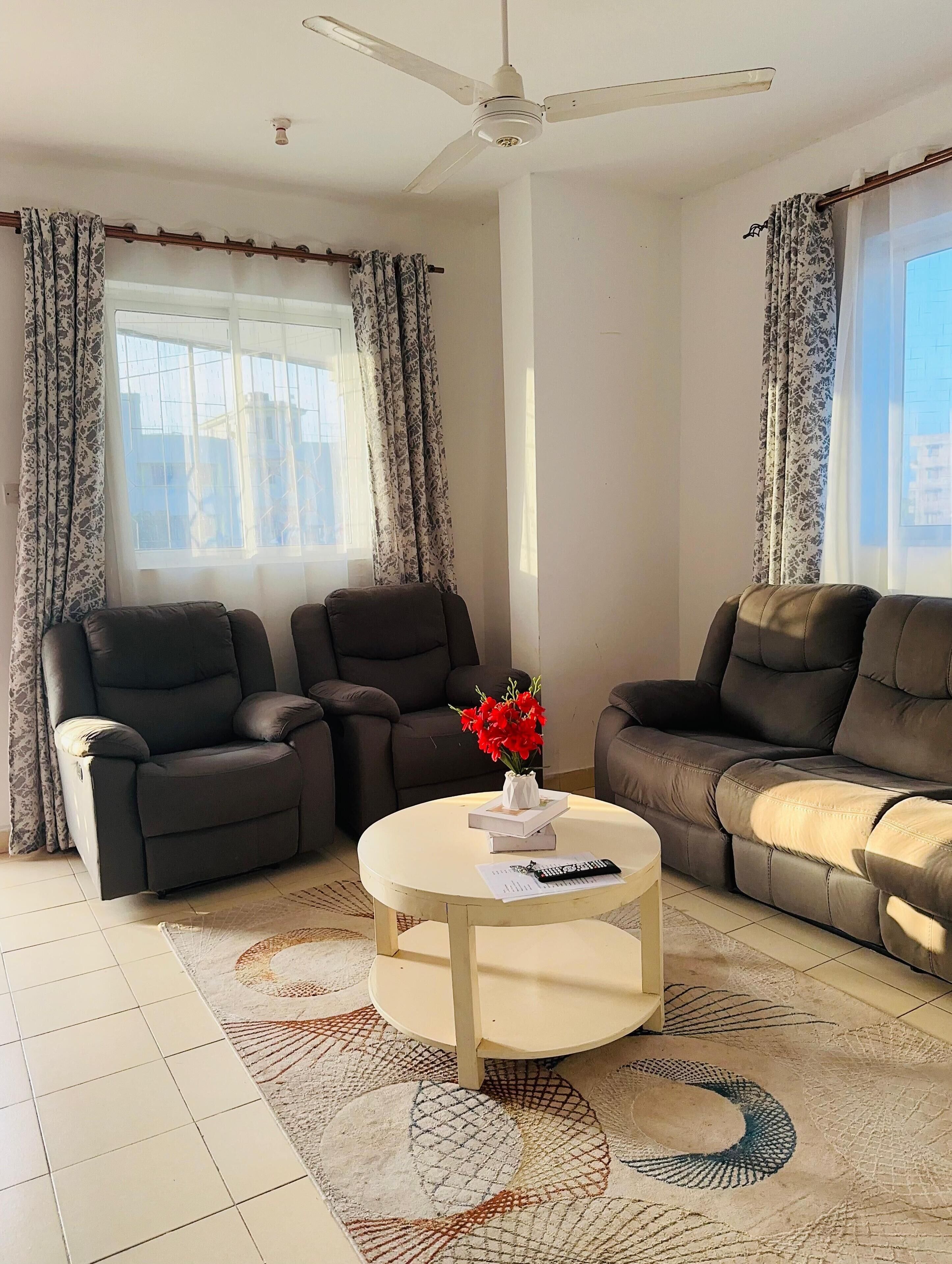 A living room with two reclining sofas, a small round coffee table with a vase of red flowers, and a ceiling fan.