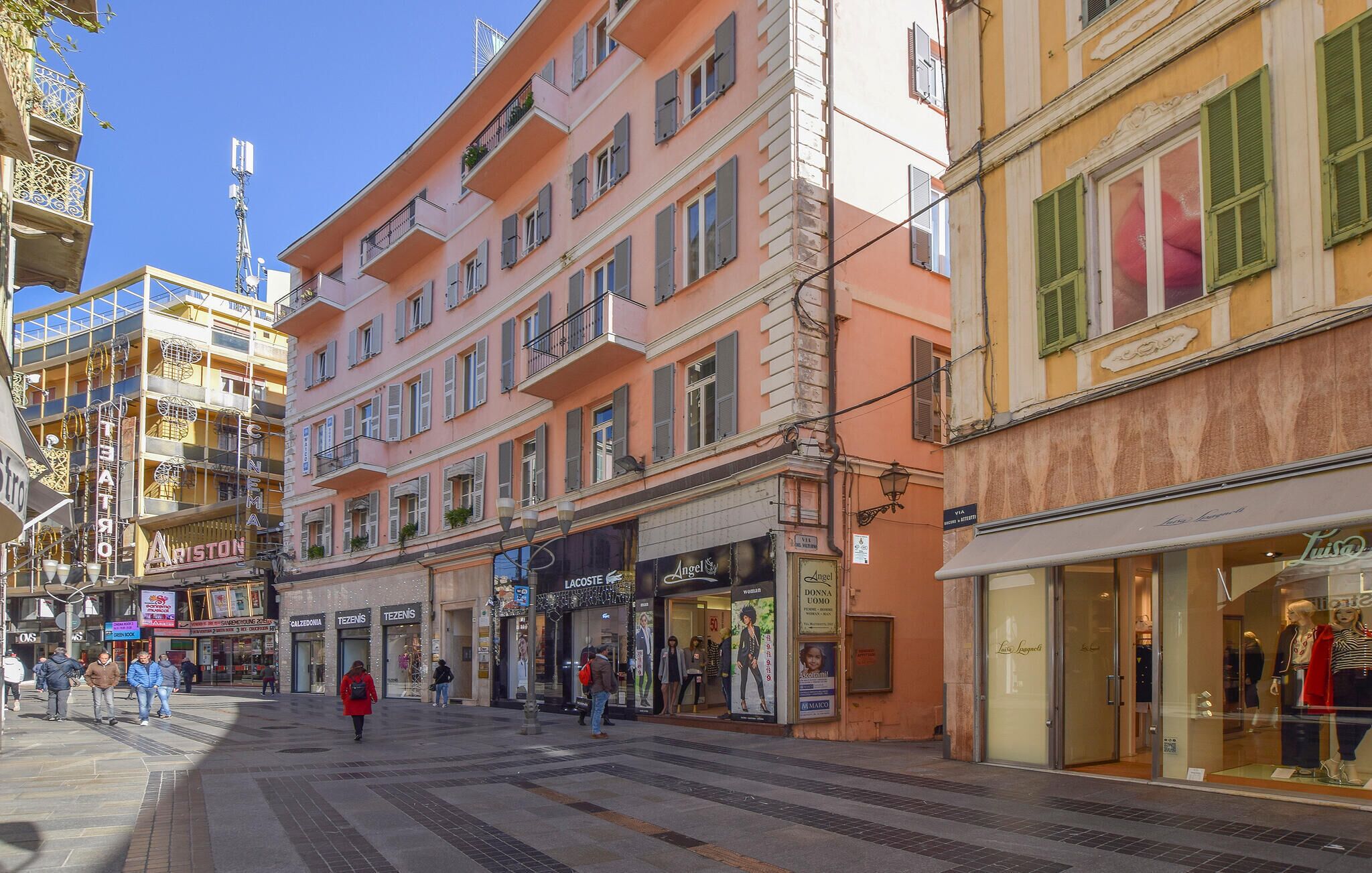 Straßenansicht mit mehrstöckigen Gebäuden, Geschäften und Fußgängern.
