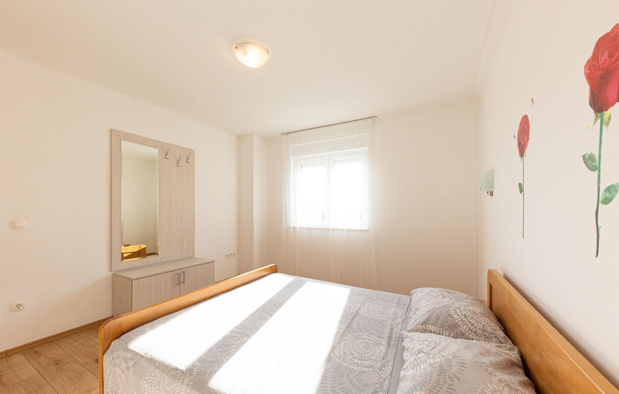 A bedroom with a bed, a mirror, a window with curtains, and a wall with a rose decoration.