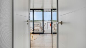 Ein modernes Zimmer mit einer großen Glasdoor, die zu einer Terrasse mit Holzmöbeln und Blick aufs Wasser führt.