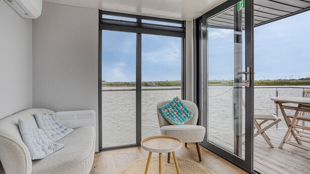 Ein modernes Zimmer mit einer großen Glasschiebetür, die auf eine Terrasse mit Blick auf einen Strand führt. Im Inneren befindet sich eine Couch mit Kissen, ein kleiner runder Tisch und ein Sessel mit gemustertem Kissen.