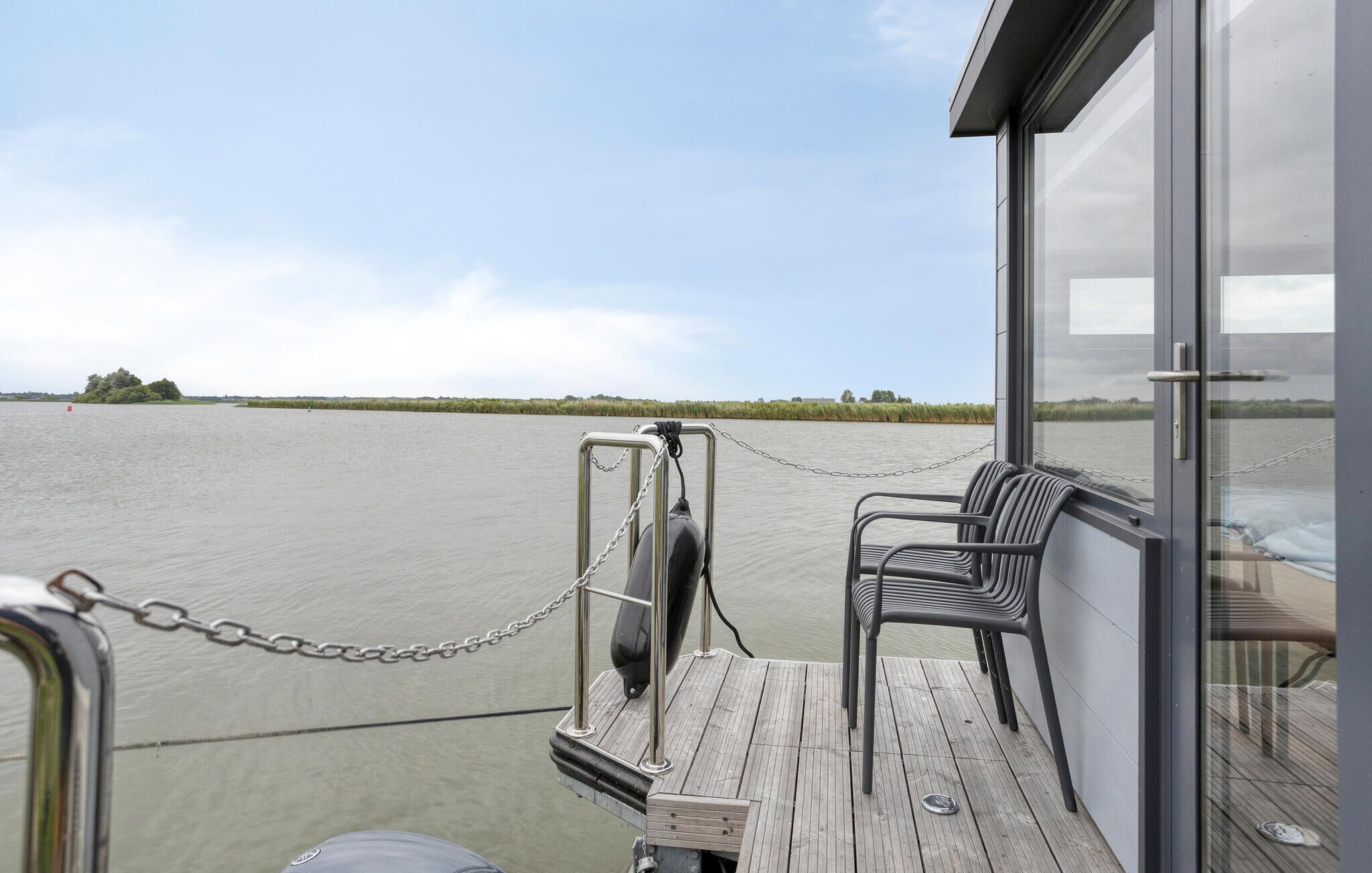 Een dek van een boot met een ketting naar een steiger, een kleine tafel en twee stoelen.