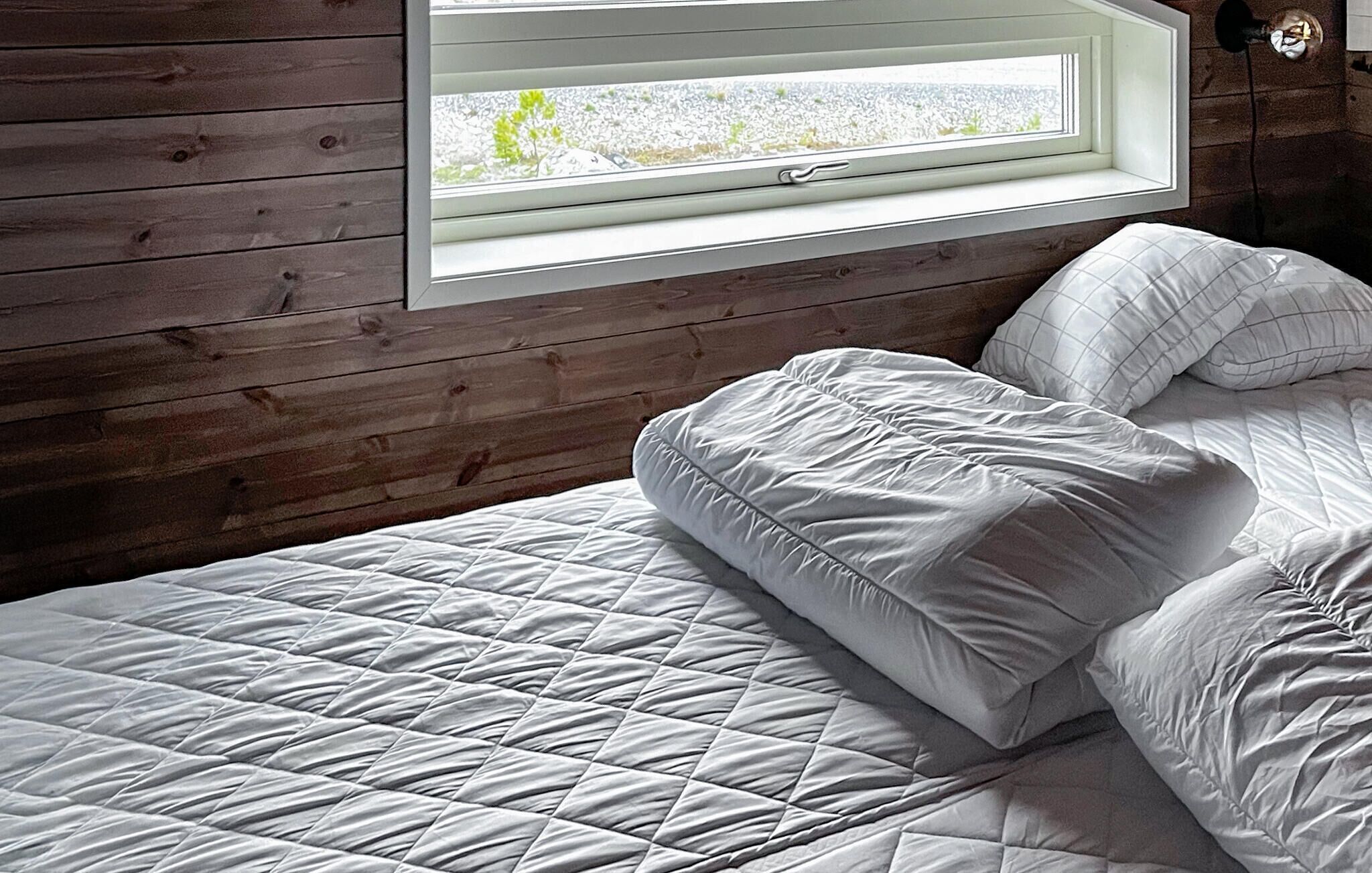 Una cama con edredón blanco acolchado y almohadas en una habitación con paredes de madera y una ventana que da a árboles.