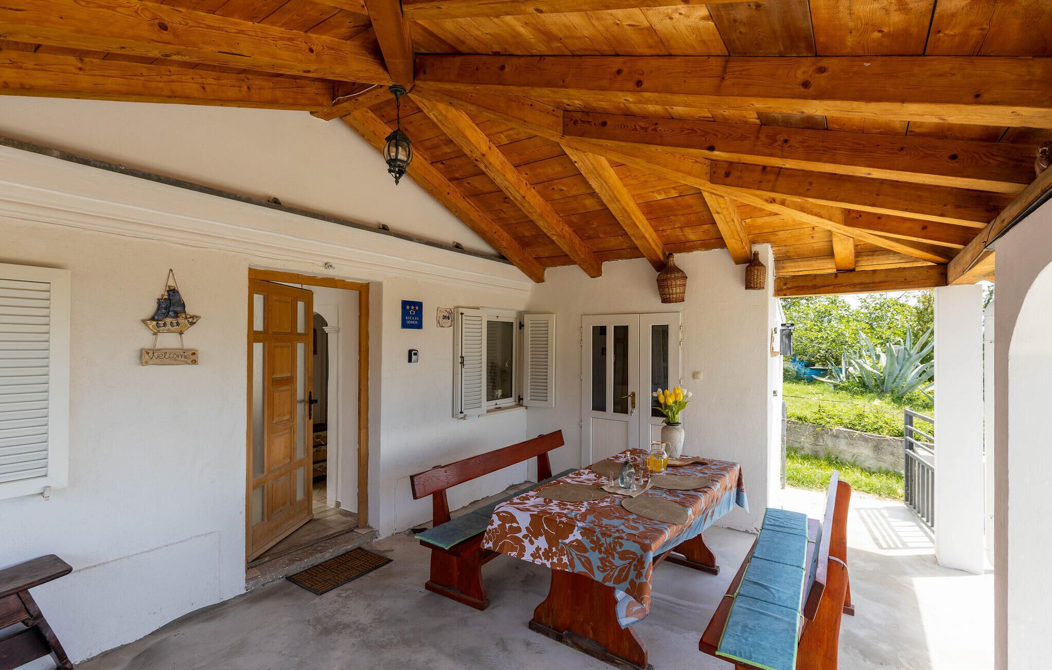A covered outdoor dining area with wooden benches and a table, a door, and a view of greenery outside.