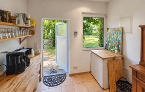Une cuisine de taille réduite, dotée d’une fenêtre, d’une étagère en bois où sont rangés les ustensiles de cuisine, et d’une porte donnant sur l’extérieur.