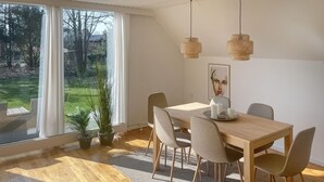 A dining area with a wooden table, four chairs, two hanging wicker lamps, a framed artwork, and a view of a garden through large windows.