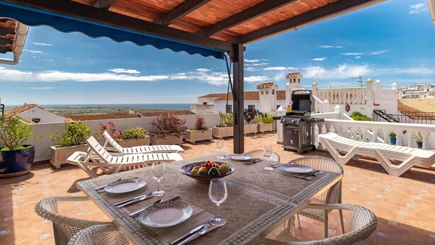 Un área de comedor al aire libre con vista al mar, una parrilla y mobiliario exterior blanco.