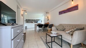 A modern living room with a sofa, a coffee table, and a TV on a cabinet. There is a kitchen in the background with white cabinets and a dining area with chairs. - Gorgeous home in Saint Pons La Calm (Saint Pons La Calm)