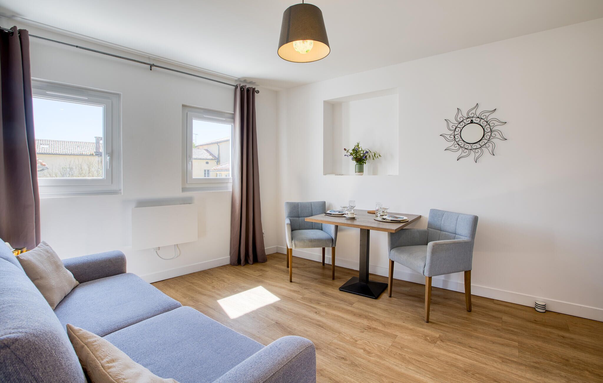Een moderne woonkamer met een grijze bank, een houten eettafel met twee stoelen, een hanglamp en een decoratief wandobject.