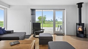 Un salon moderne avec un canapé, un pouf et une table basse en bois. Une chaise noire est placée près d’une porte-fenêtre coulissante qui donne sur une terrasse extérieure. Un poêle à bois noir est visible dans un coin.