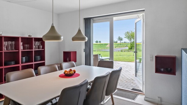 Un coin repas avec une table blanche, des étagères rouges et une vue sur un paysage verdoyant, visible à travers une porte vitrée.
