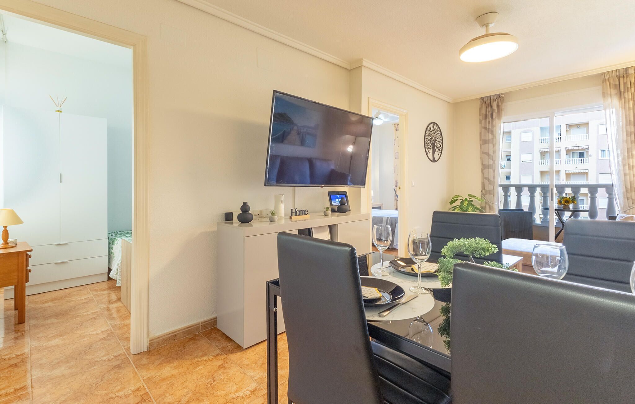 A dining area with a TV mounted on the wall, a small table with chairs, and a balcony with a view of buildings.