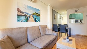 Un salon avec un canapé beige, une table à manger avec des chaises, un coin cuisine avec des meubles de couleur turquoise et un tableau représentant un paysage côtier.