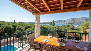 A terrace with a wooden table set for dining, overlooking a pool and a scenic view of a lake and mountains.