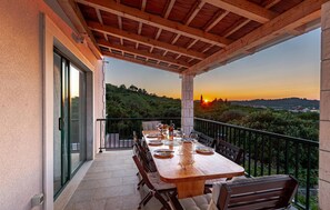 A wooden table set for dining on a balcony with a view of a sunset over a hilly landscape. - Beautiful home in Pupnat (Pupnat)