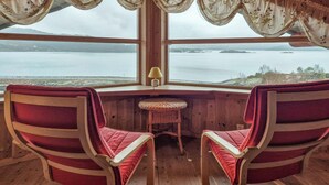 Eine Hütte mit zwei roten Polstersesseln, einem kleinen Korbstuhl und einer Lampe. Ein großes Fenster bietet Blick auf einen See und entfernte Ufer.