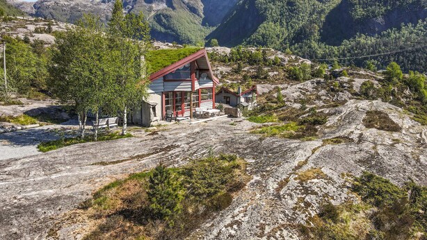 Eine rustikale Holzhütte mit rotem Dach und weißen Wänden, umgeben von felsigem Gelände und dichtem Wald.