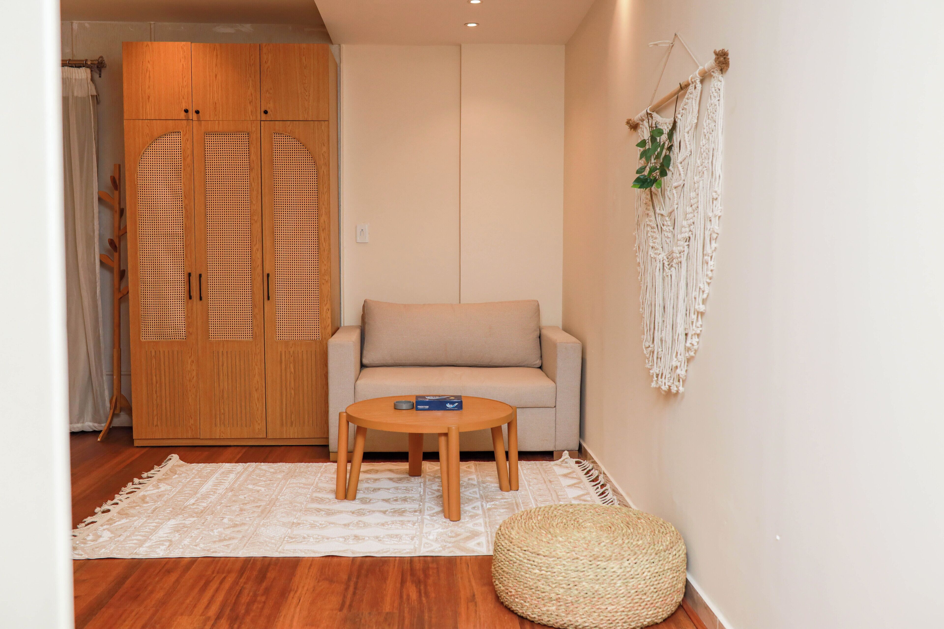 A room with a wooden wardrobe, a beige sofa, a small round table, and a macrame wall hanging.
