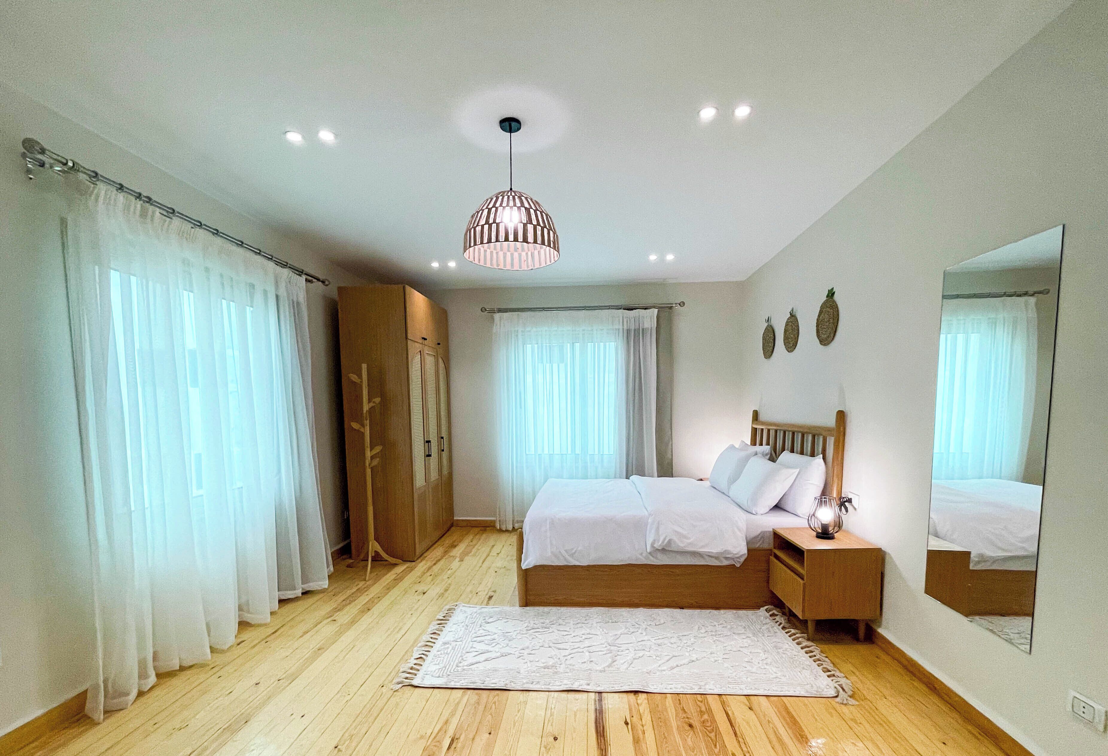 Ein Schlafzimmer mit Holzboden, einem Bett mit weißer Bettwäsche, einem Nachttisch, einem Spiegel und einer Deckenleuchte.