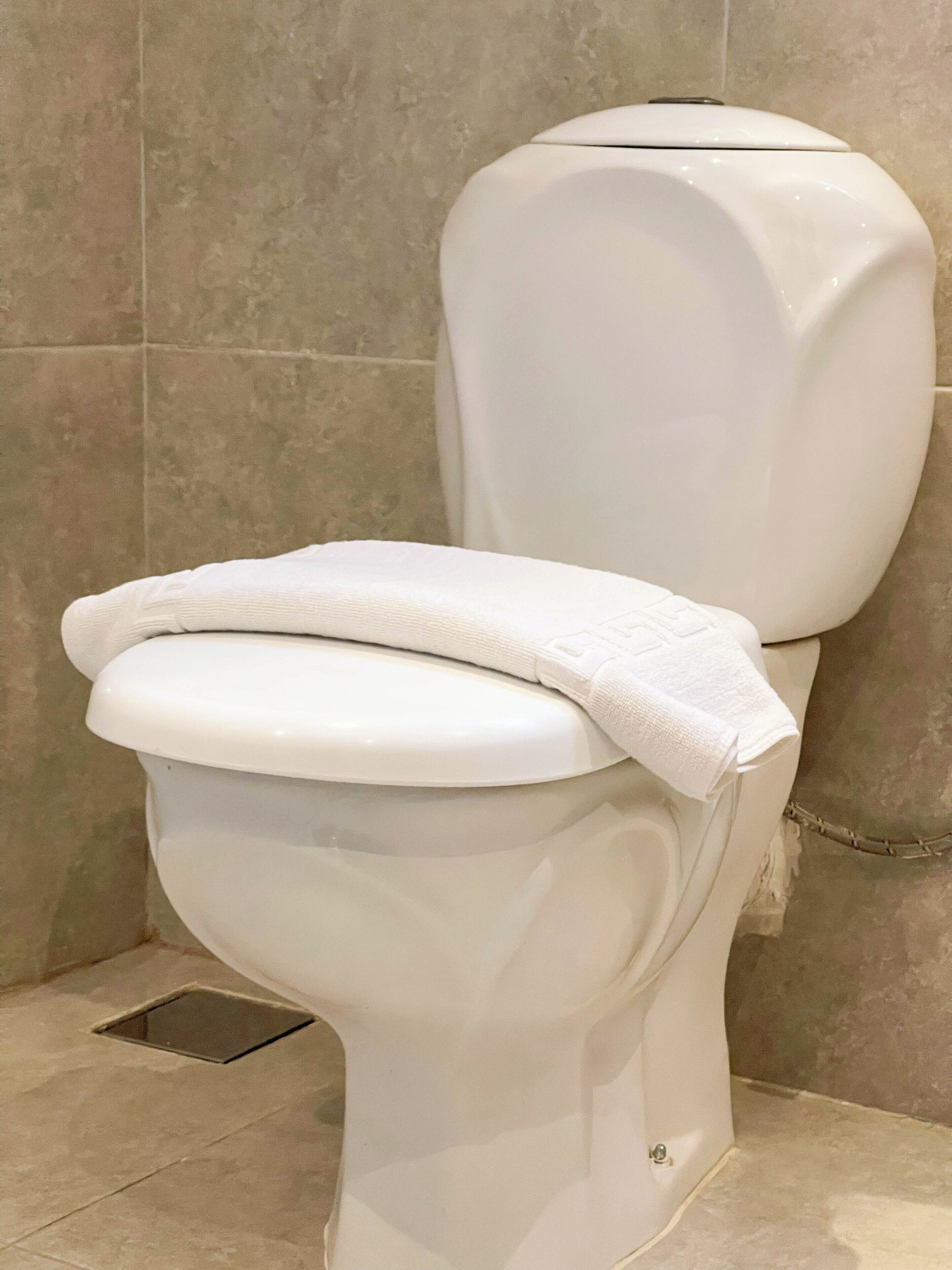 Sanitário com vaso sanitário branco e toalha no assento, revestido com azulejos bege.