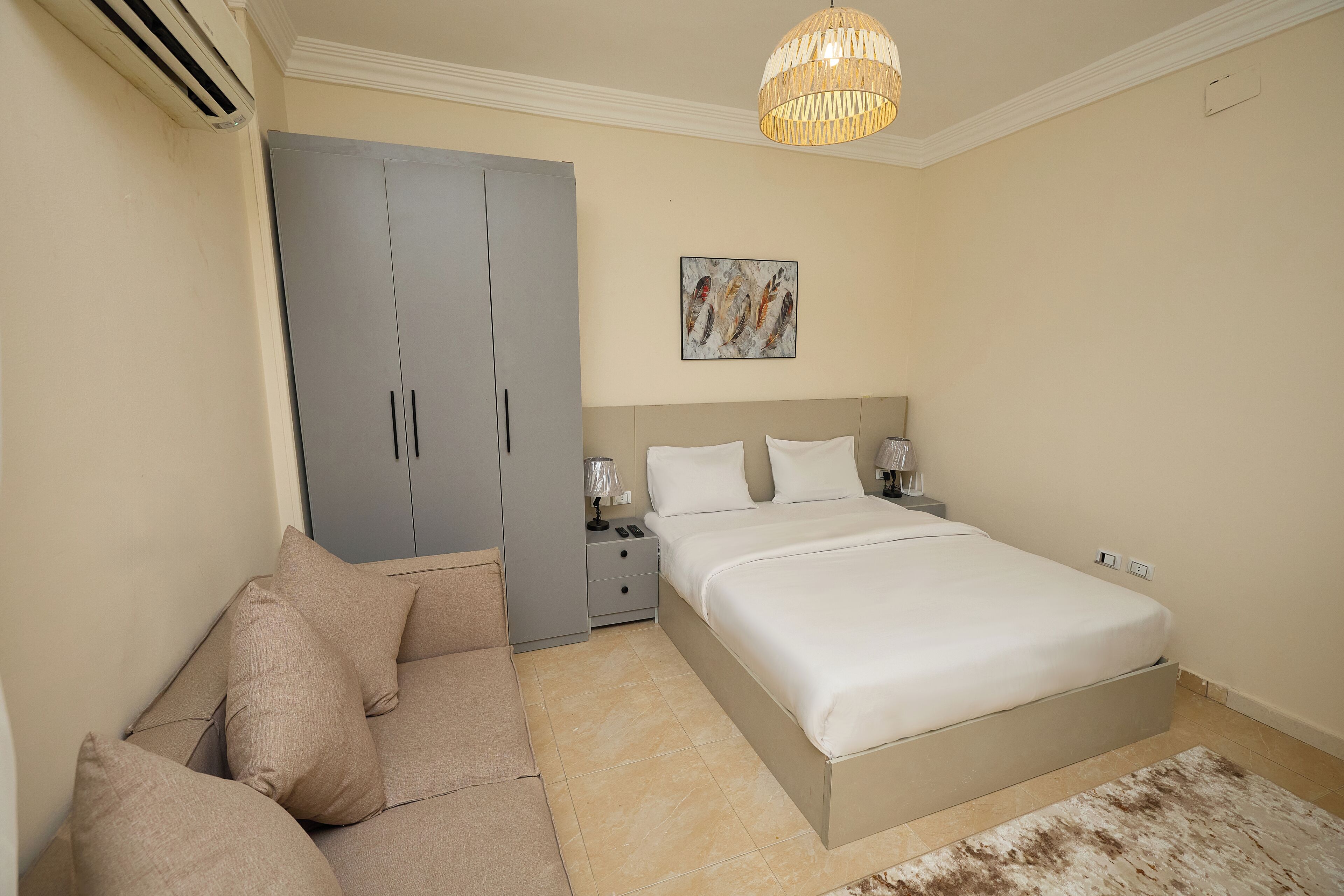 A bedroom with a bed, a sofa, a wardrobe, a nightstand, a lamp, and a ceiling light fixture.