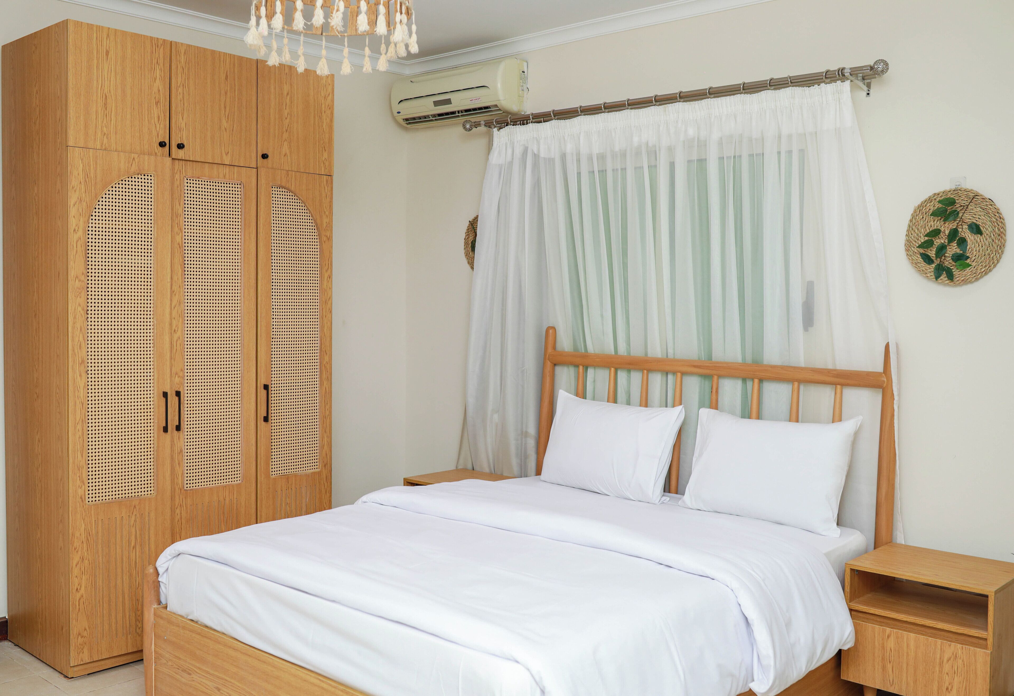 Un dormitorio con cama de madera, ropa de cama blanca, armario de madera con puertas persianas, mesita de noche y aire acondicionado de pared.