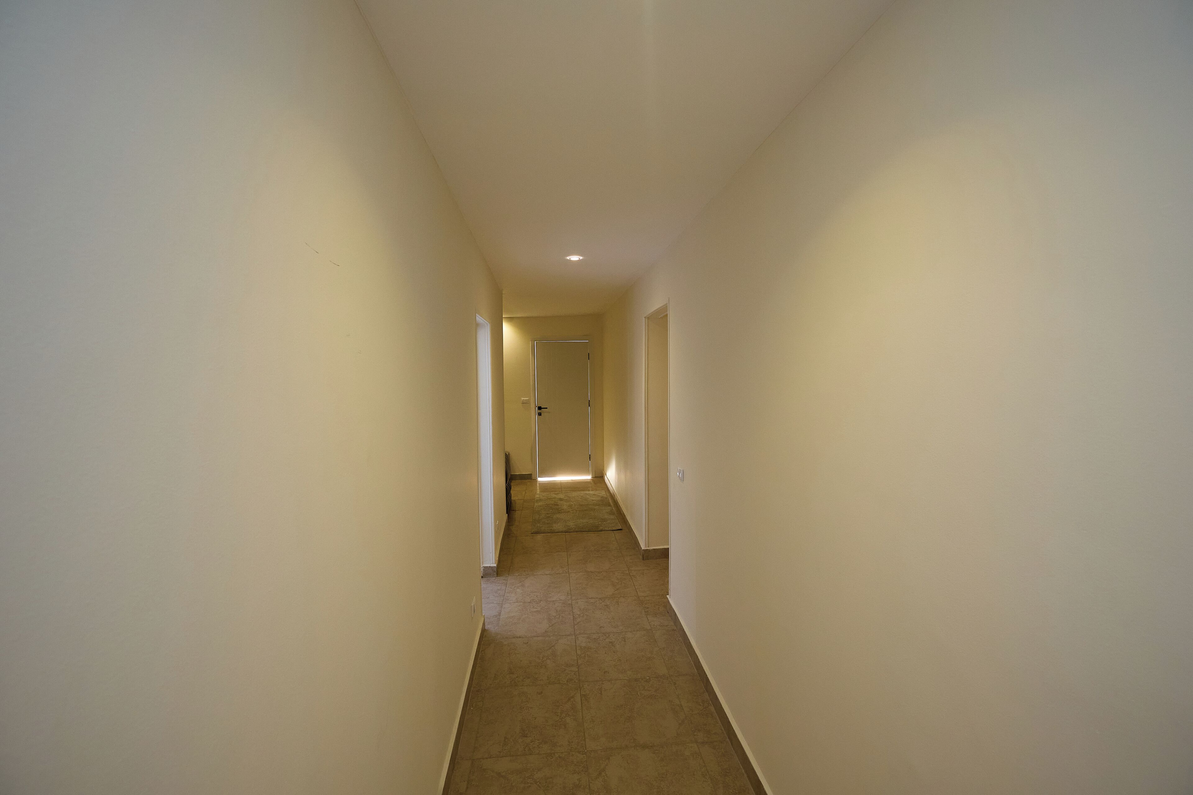 A narrow, well-lit hallway with a carpeted floor and plain walls, leading to a door at the end.