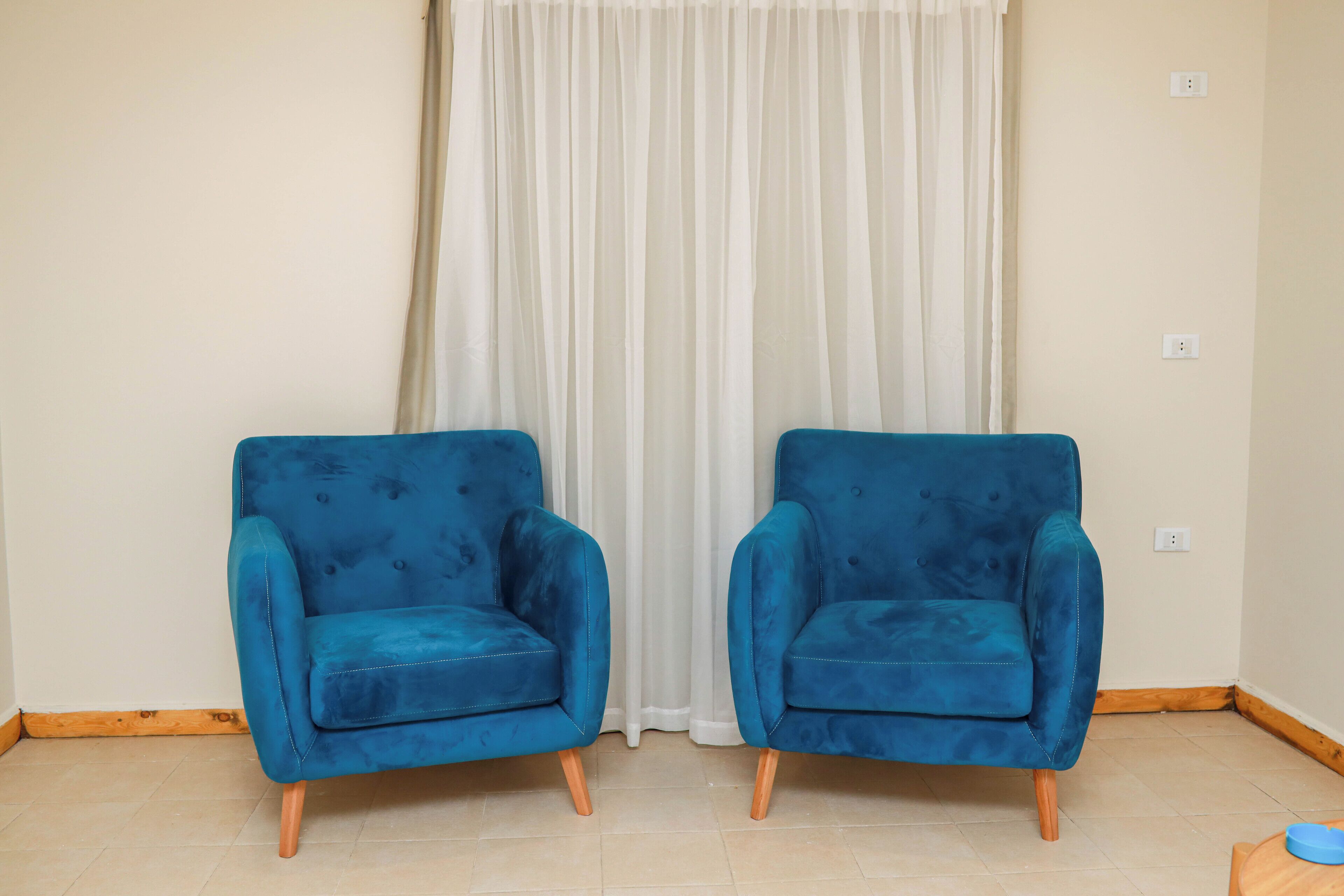 Dos sillones azules con patas de madera en una habitación con cortina blanca y paredes de tonos claros.