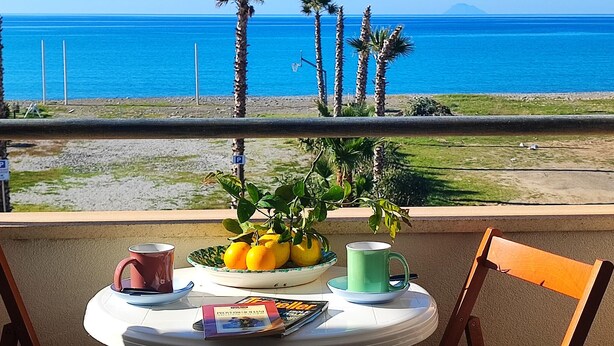 Une table avec un saladier rempli de citrons et d’oranges, deux tasses et un magazine, le tout placé sur un balcon donnant sur une plage bordée de palmiers et sur l’océan.