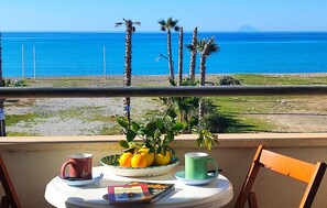 Una mesa con un bol de limones y naranjas, dos tazas y una revista en un balcón con vistas a una playa con palmeras y el océano.