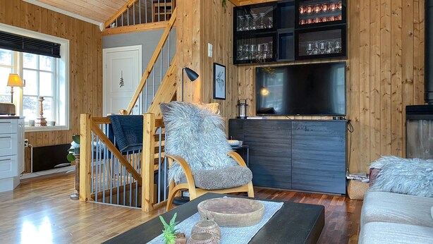 A cozy living room with a wooden staircase, a comfortable chair, a flat-screen TV, and a glass cabinet with decorative items.