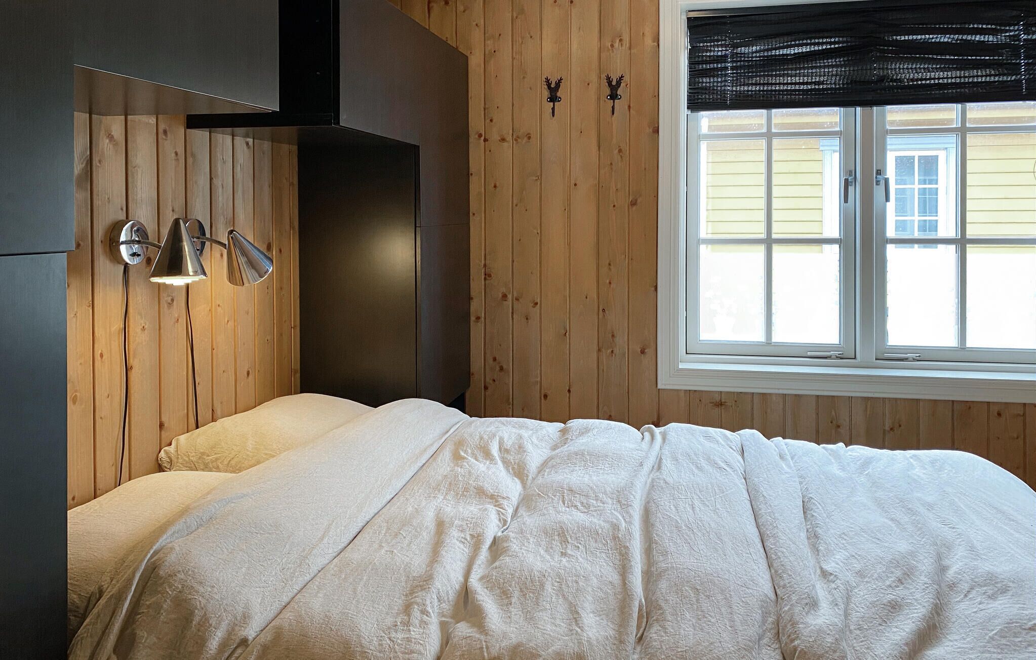 Un dormitorio con cama, paredes de paneles de madera, ventana con persianas y una lámpara fijada a la pared.