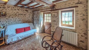 Una habitación rústica con pared de piedra, techo de madera, sofá blanco con cojines rojos, una silla de mimbre y una ventana con vistas.