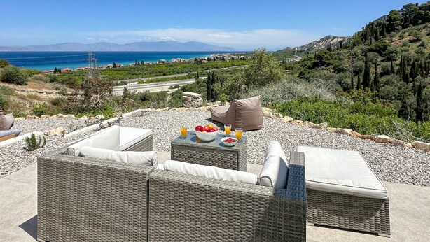 Una terraza con mobiliario de mimbre, una mesa con fruta y bebidas, y una vista panorámica del mar y las colinas.