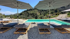 Zona junto a la piscina con sillones de descanso y sombrillas, rodeada de suelo cubierto de guijarros y un paisaje montañoso.