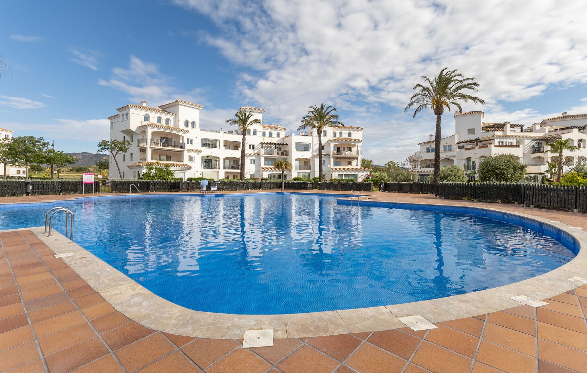 A swimming pool with a tiled edge, surrounded by a fence, and adjacent to a multi-story building with balconies and palm trees.