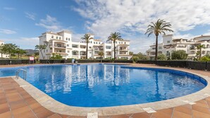 Una piscina con borde de azulejos, rodeada por una valla, junto a un edificio de varios pisos con balcones y palmeras.