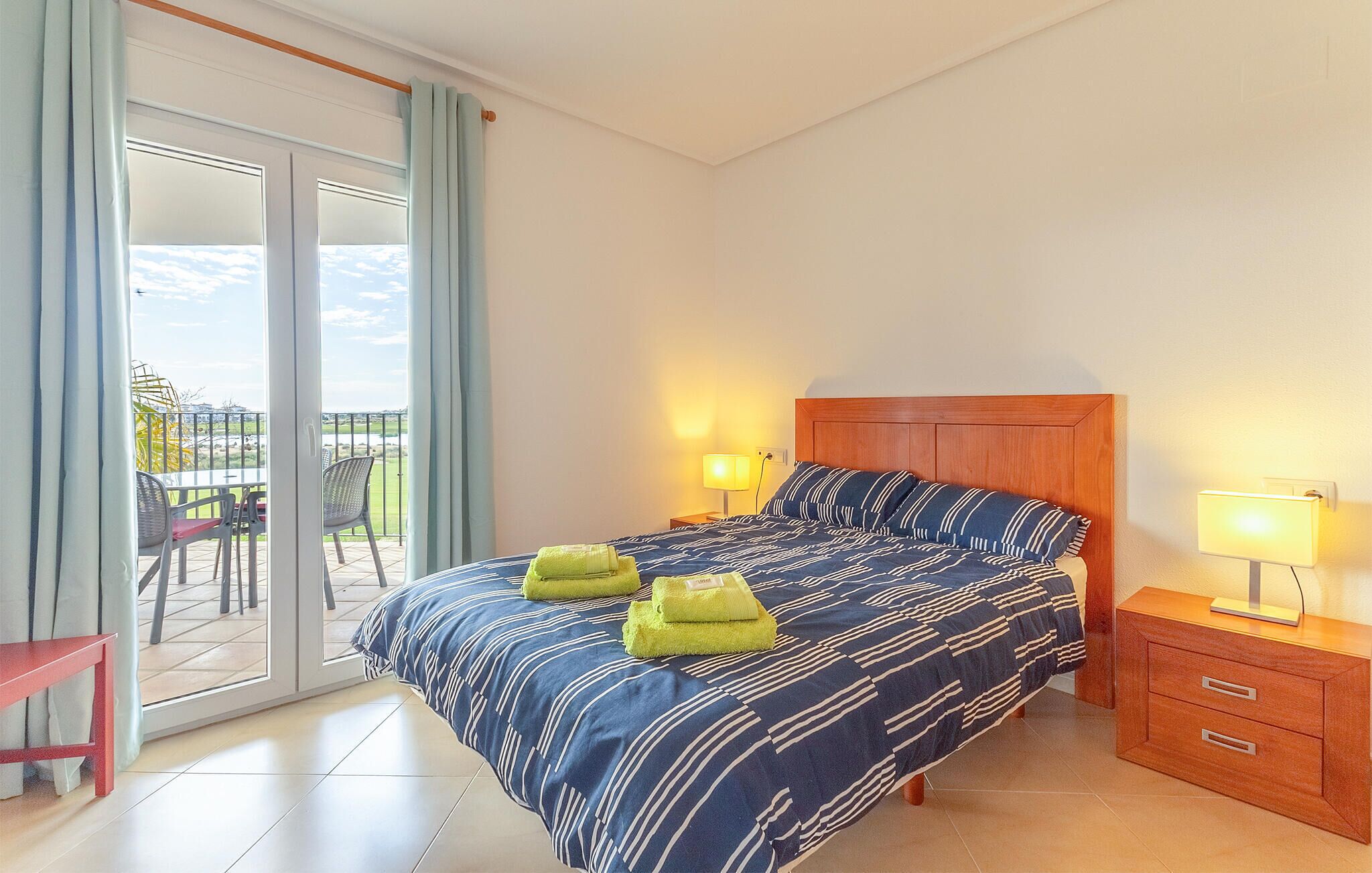 A bedroom with a bed, two bedside lamps, a wooden nightstand, and a view of the outside balcony.