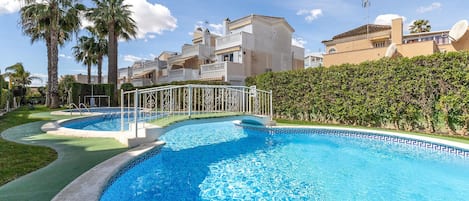 A swimming pool with a geometric tile pattern, surrounded by a white metal fence, palm trees, and a hedge.