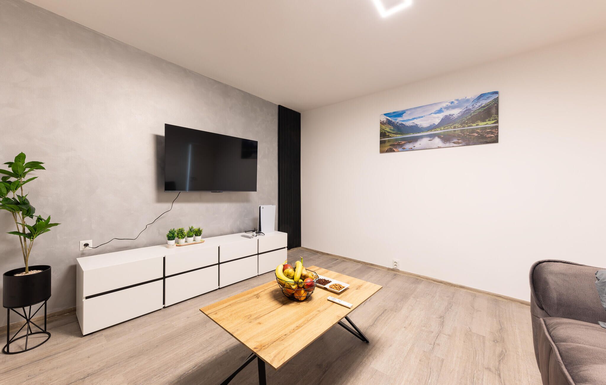 Una sala de estar moderna con un televisor de pantalla plana montado en la pared, una mesa de centro de madera con frutas y aperitivos, un sofá gris y una planta en maceta.