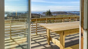Een houten terras met een tafel en banken, met uitzicht op een kustlandschap met heuvels en de zee.