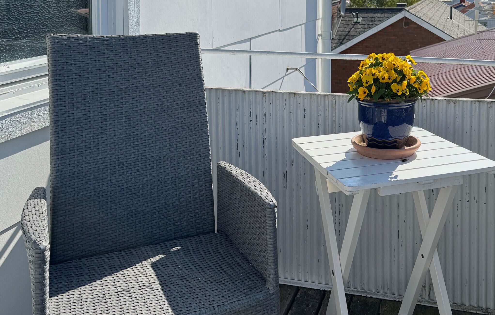 A balcony with a wicker chair and a small white table with a blue pot of yellow flowers.