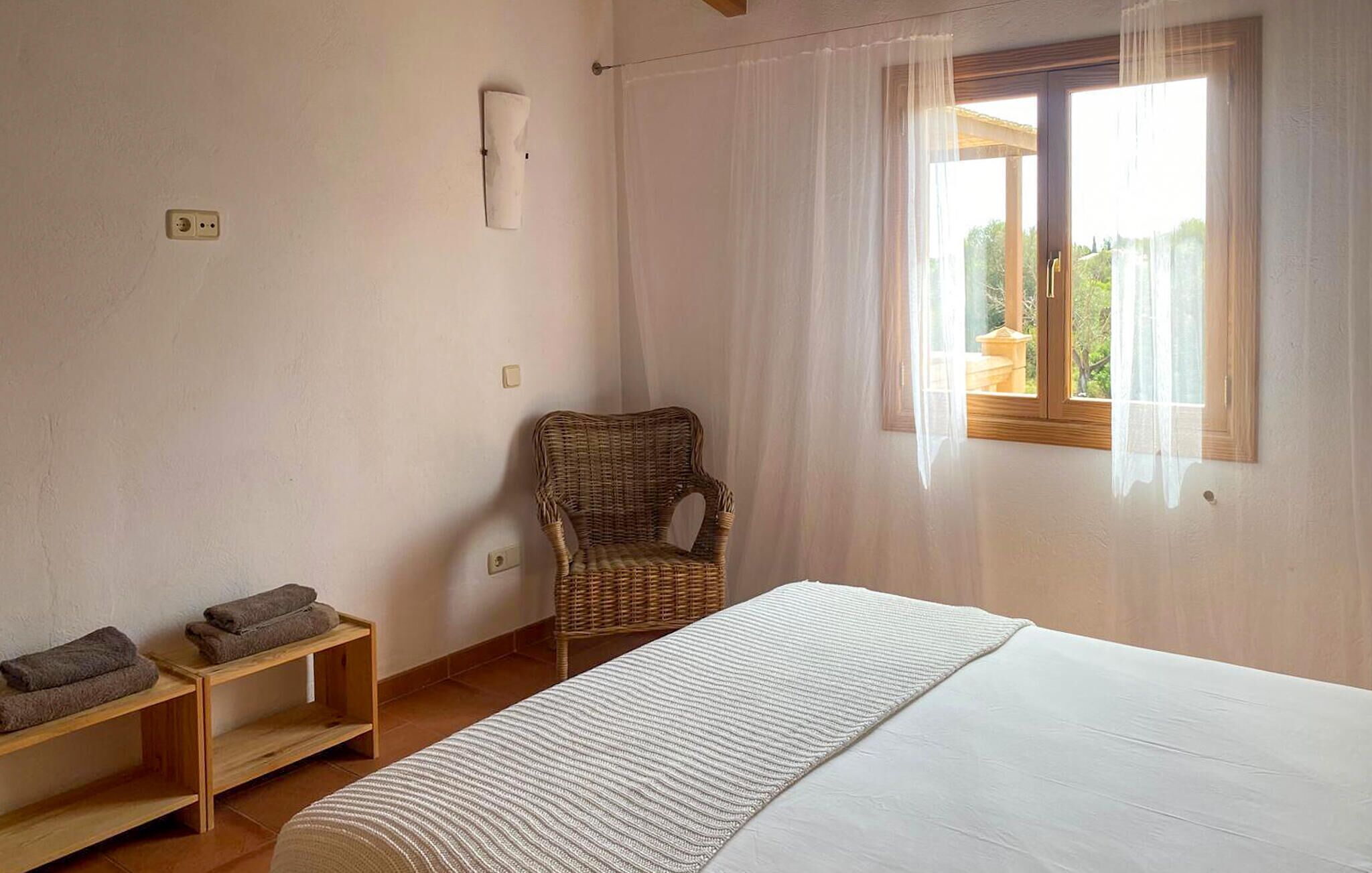 Une chambre à coucher comprenant un lit, une chaise, une étagère avec des serviettes, une fenêtre avec des rideaux et un luminaire fixé au mur.