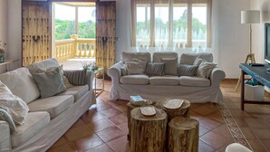 A living room with a beige sofa, a white sofa, and a wooden coffee table. There is a balcony with a wooden railing and a view of greenery. - Nice home in Son Servera (Son Servera)