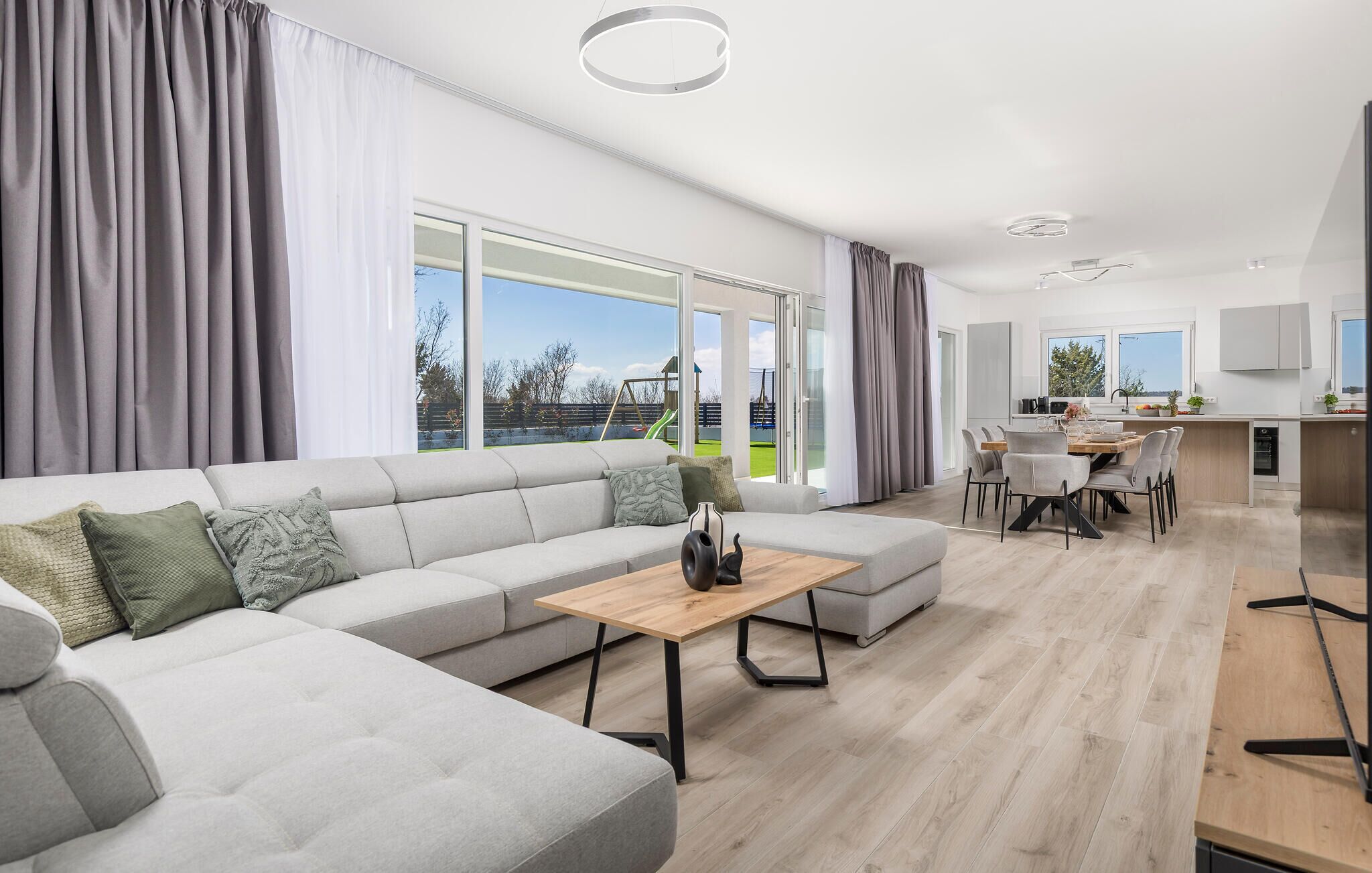 A modern living room with a grey sectional sofa, a wooden coffee table, and a dining area with a wooden table and chairs.