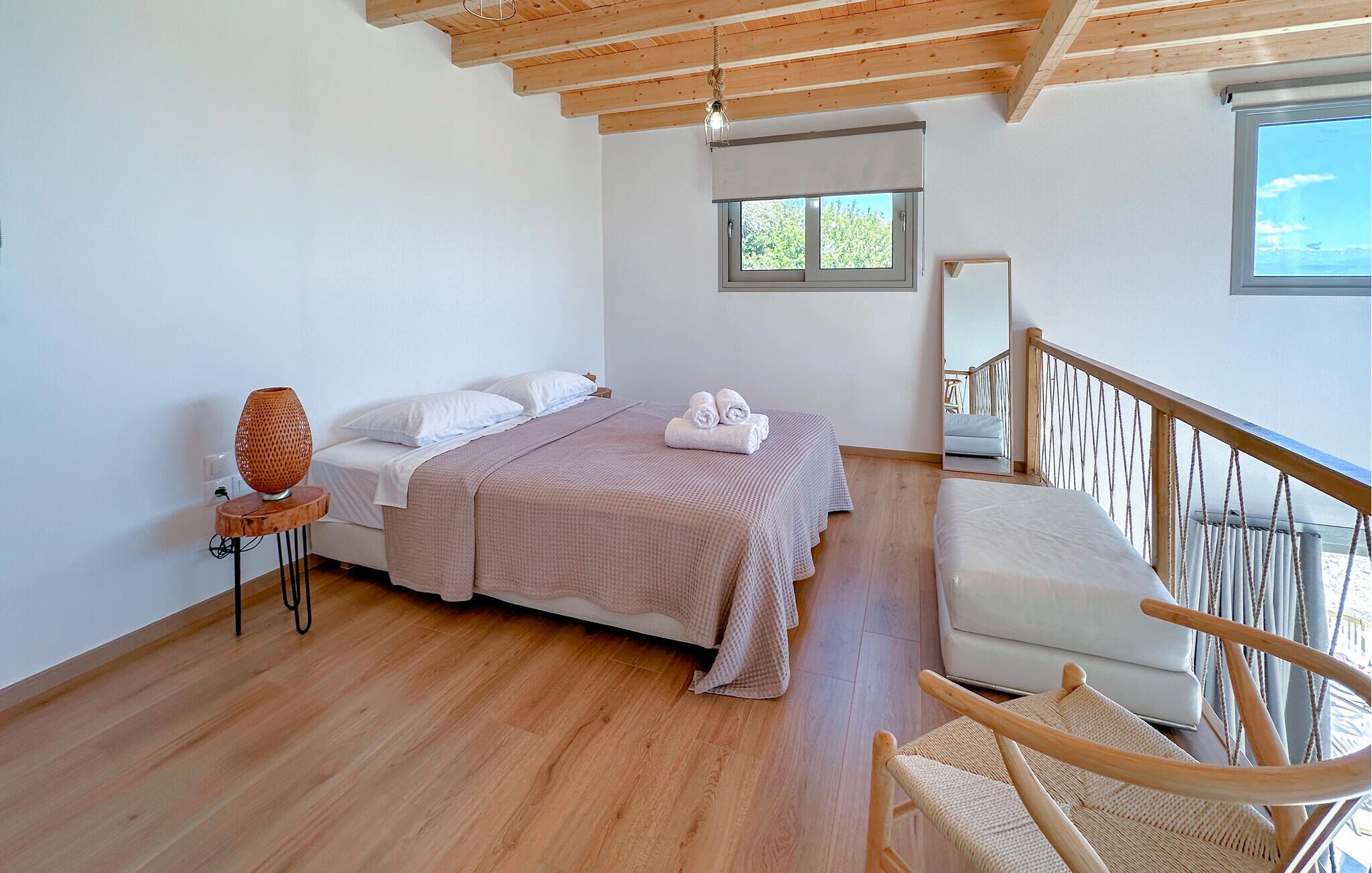 A bedroom with a bed, a chair, a small table, a window, and a balcony railing.