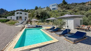 Un área de piscina moderna con sillas de descanso y sombrillas, bordeada por una playa de guijarros y una colina con edificios.