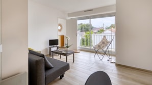 A modern living room with a grey sofa, a small table, a TV, and a decorative mirror. A chair is placed near a large window with a view of a balcony and buildings.