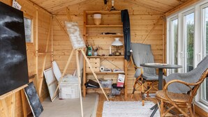 A cozy wooden cabin interior with a painting easel, a wicker chair, a small table, and a window providing a view of greenery.